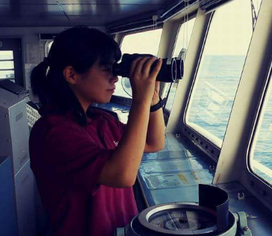 Lookout duties: A Seawoman on the ship's bridge holding a binocular and observing the horizon.